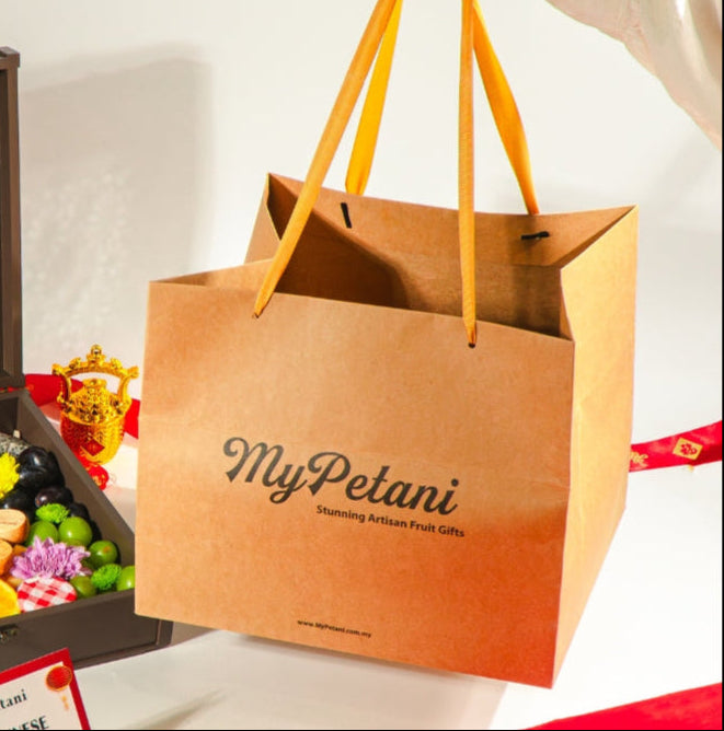 A person holding a beige carry-gift bag with the text 'mypetani' printed on it, alongside a wooden gift box and a variety of fruits.