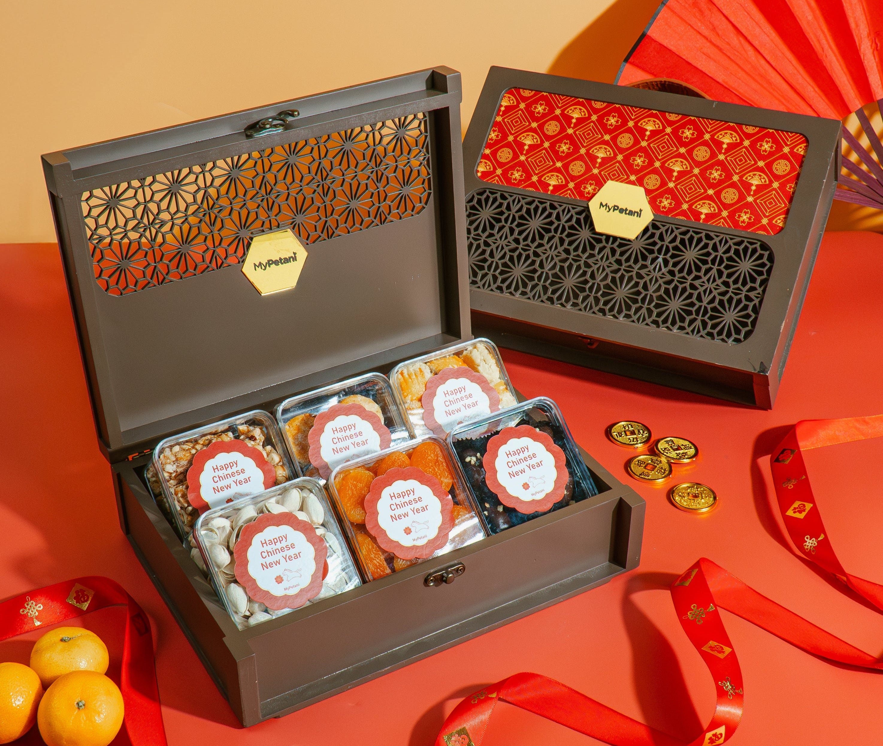 Decorative Chinese New Year gift box with snacks on a red surface, featuring a red fan and tangerines.
