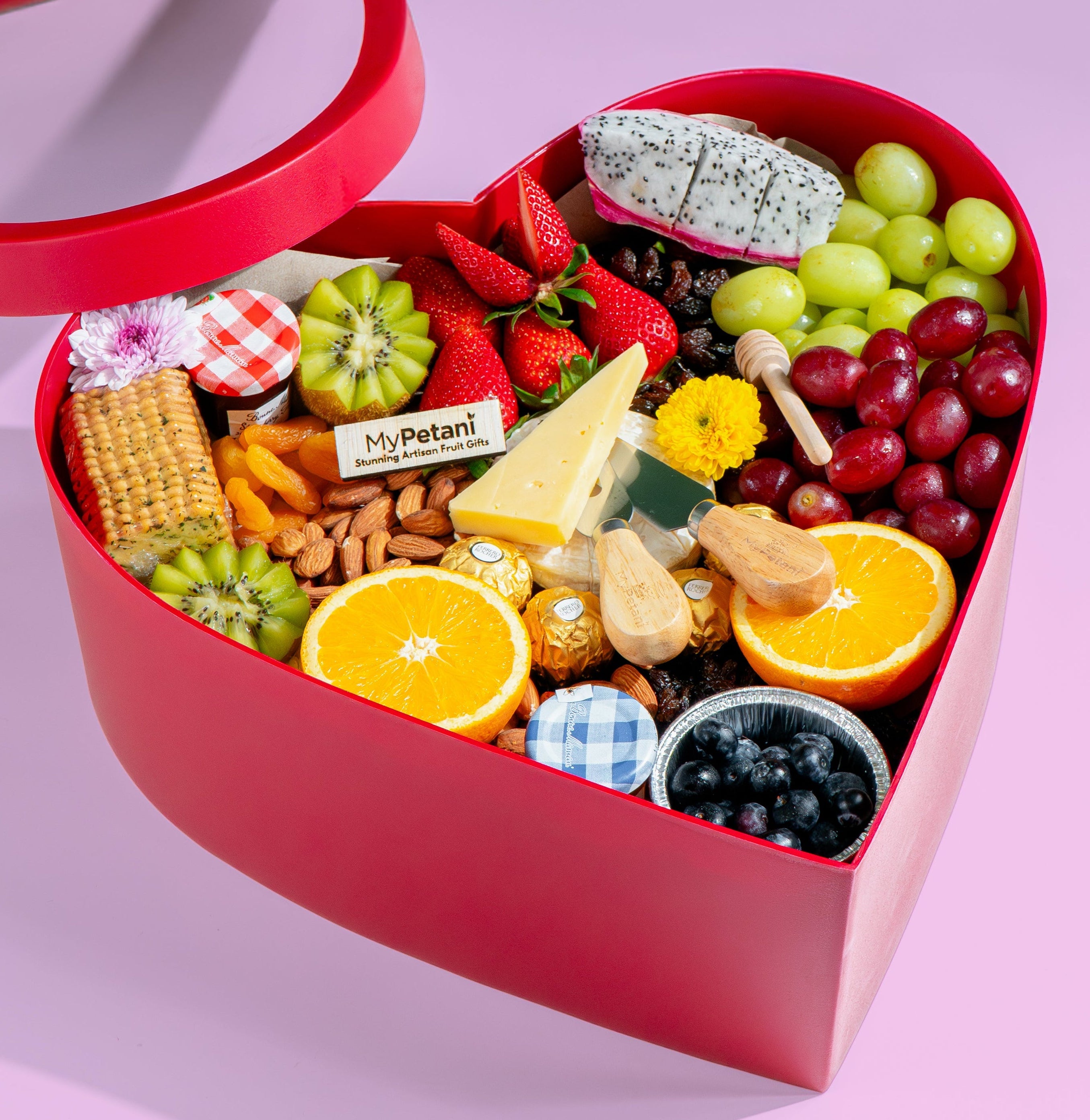 Heart-shaped box filled with fruits, snacks, and treats on a pink background