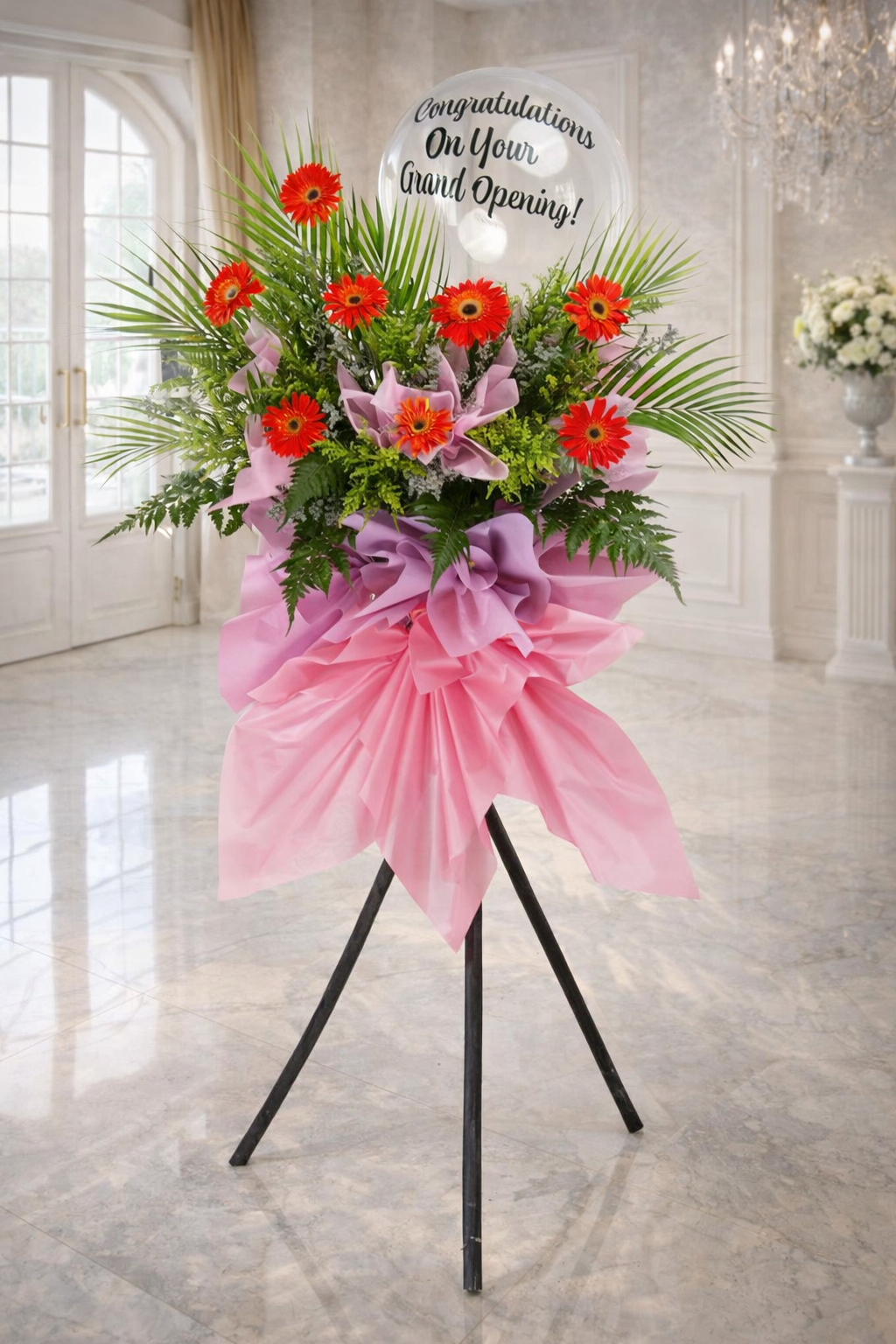 Floral arrangement with red flowers and pink ribbon on a stand in a room with large windows.