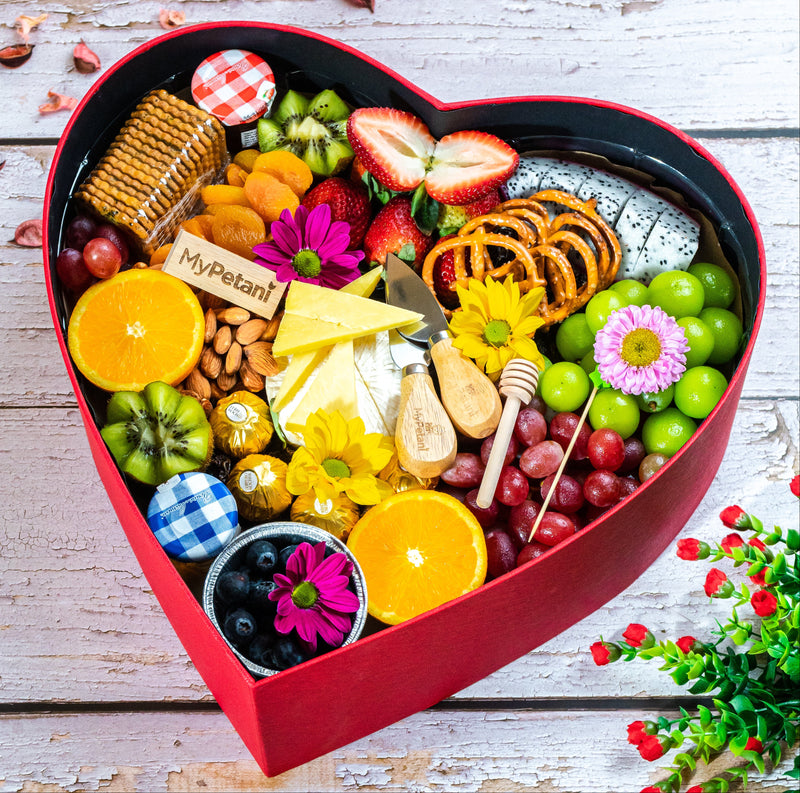 A heart-shaped red cheese grazing box containing an assortment of fruits, cheeses, nuts, and sweet items, presented as a gift.
