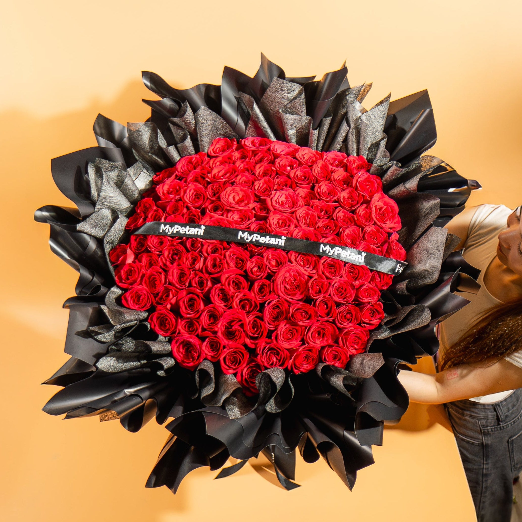 Heart-shaped arrangement of red roses with black leaves held by a person against an orange background
