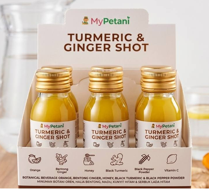 Three bottles of MyPetani Turmeric & Ginger Shot arranged in a retail display box on a wooden surface, with a blurred kitchen background showing a glass and honey dipper.